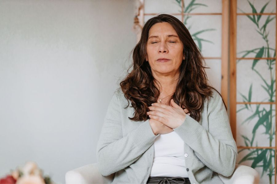 Eine Frau sitzt mit auf einem Sessel. Im Vorgespräch ihrer Rebalancing Sitzung hat sie die Augen geschlossen, die Hände übereinander auf ihre Brust gelegt und spürt in ihren Körper, was er ihr mitteilen möchte.